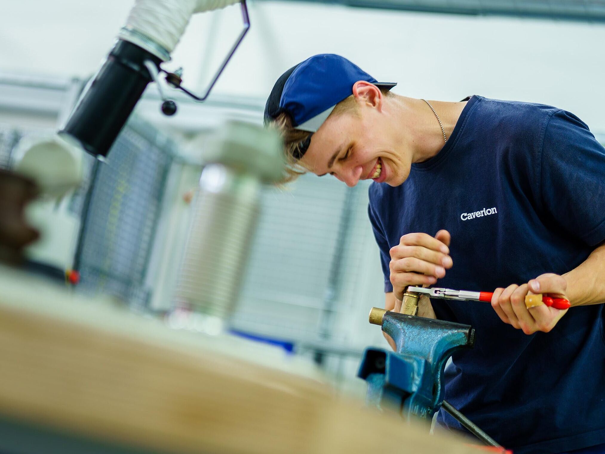 In einer Werkstatt an einer Werkbank arbeitet eine Person mit einer Rohrzange an einem Werkstück, welches in einem Schraubstock eingespannt ist.