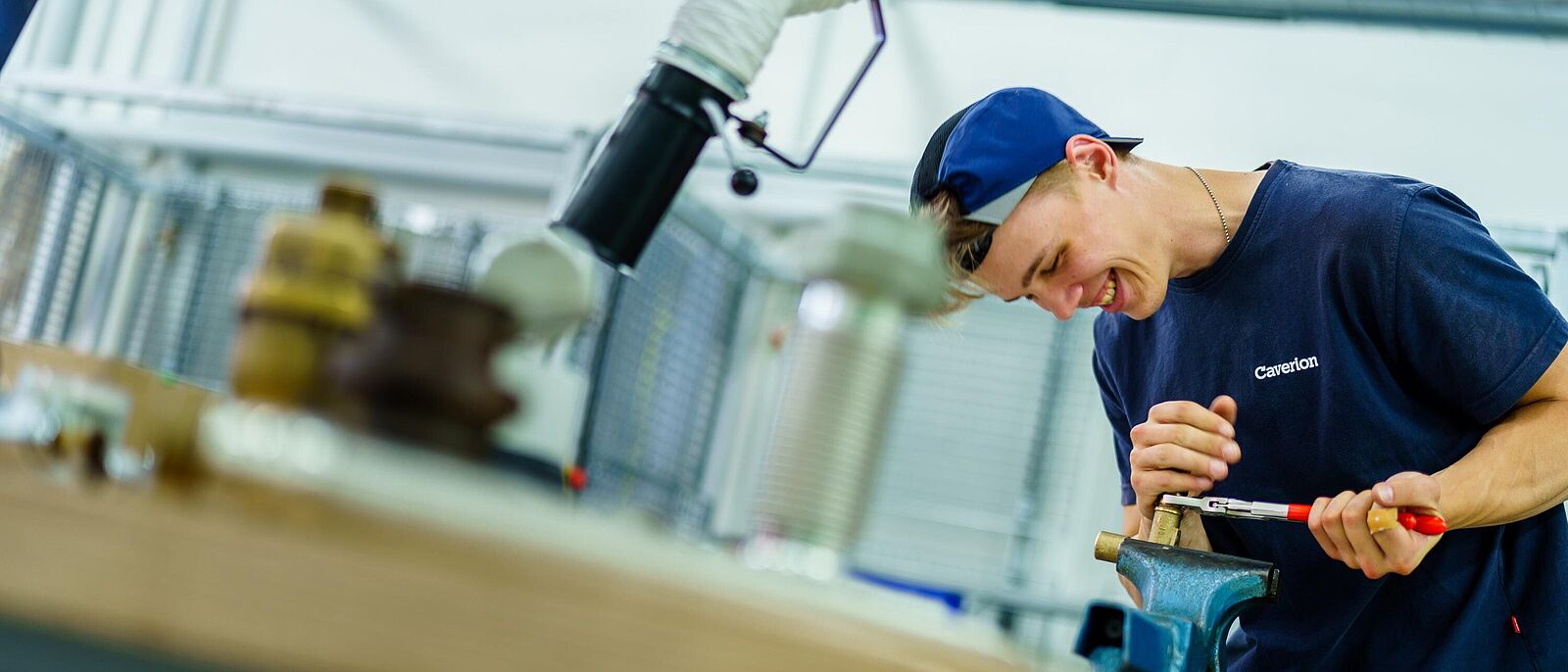 In einer Werkstatt an einer Werkbank arbeitet eine Person mit einer Rohrzange an einem Werkstück, welches in einem Schraubstock eingespannt ist.