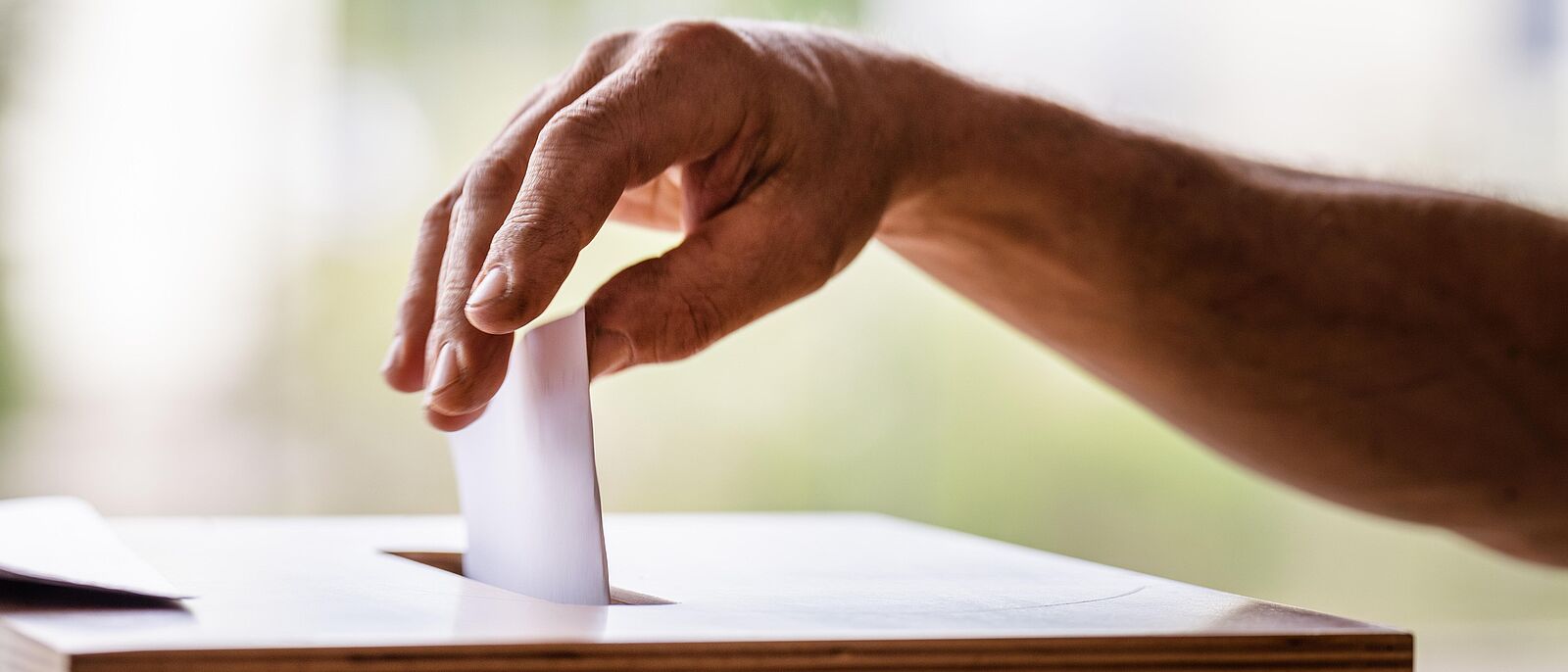 Makroaufnahme einer Hand die gerade einen gefalteten Wahlzettel in den Spalt der Wahlurne einwirft.