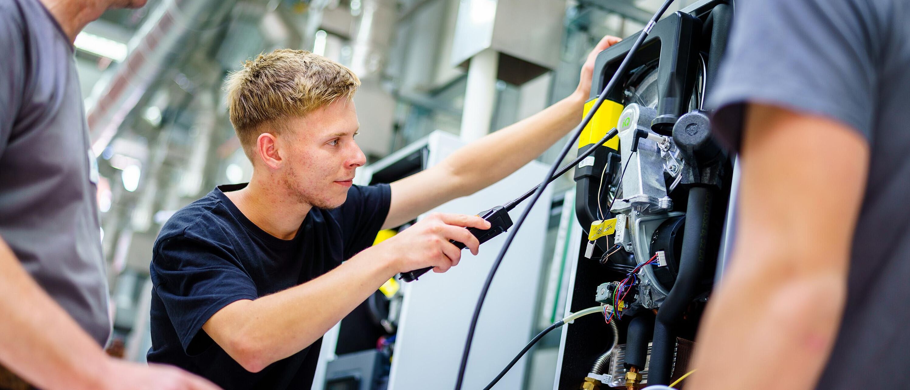 Ein Lehrling führt Arbeiten/Messungen an einem Motor ein einem Montageschrank aus und wird von zwei Ausbildern links und rechts beraten, begleitet