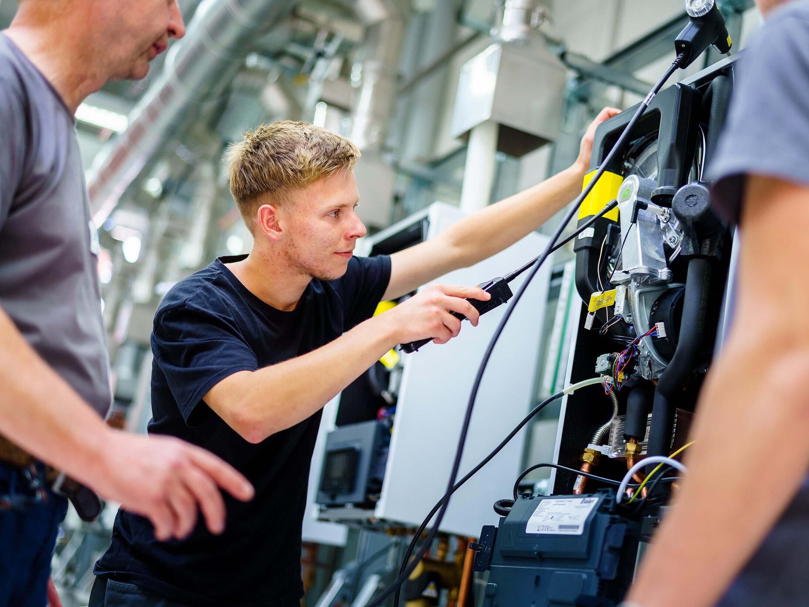 Ein Lehrling führt Arbeiten/Messungen an einem Motor ein einem Montageschrank aus und wird von zwei Ausbildern links und rechts beraten, begleitet