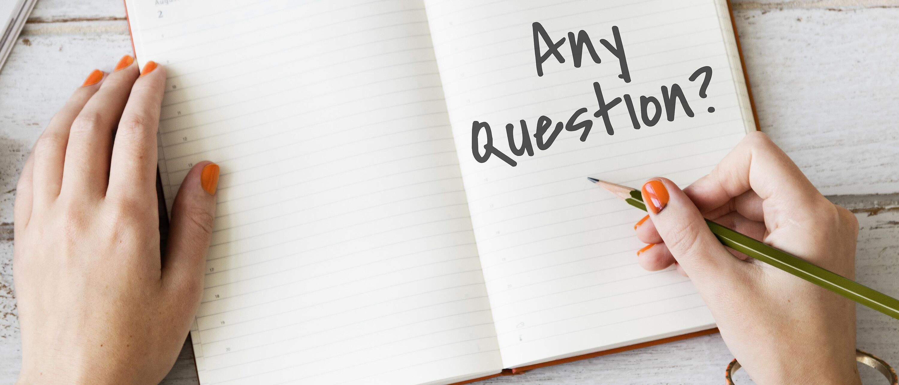 Eine Person sitz am Schreibtisch vor einem geöffneten Notizbuch und schreib mit einem Bleistift \"Any Question\" .