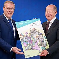 links steht der Präsident der Handwerkskammer Dresden zusammen mit Bundeskanzler Olaf Scholz halten sie ein Plakat der Karikaturaustellung