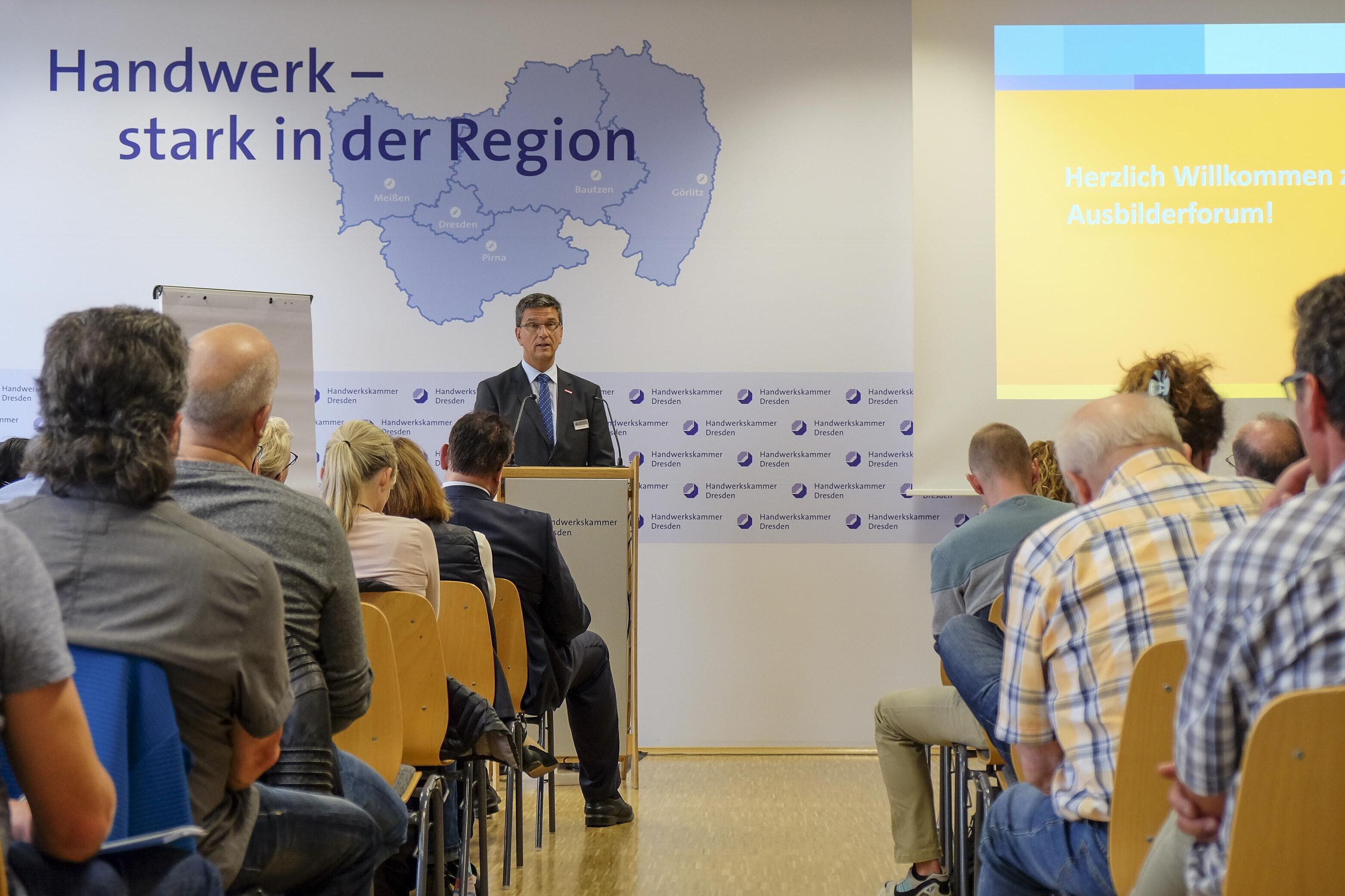 https://www.hwk-dresden.de/veranstaltungen/detail/10-ausbilderforum-2025-ausbilden-auf-augenhoehe.html Blick in dem Konferenzraum über die Zuhörer Richtung Redner. Hinter ihm eine Präsentationswand.
