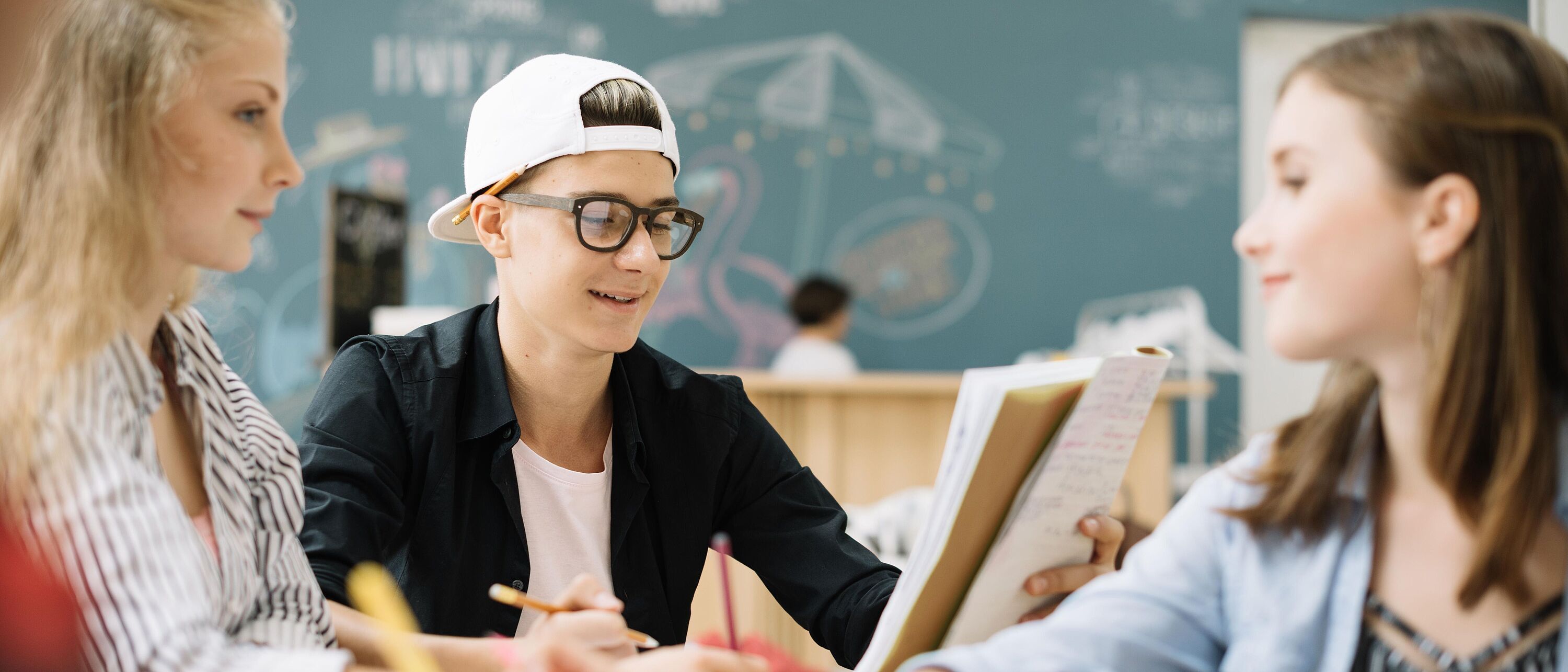 § Schüler sitzen gemeinsam am Tisch und lösen Aufgaben im Hintergrund eine Kreidetafel.