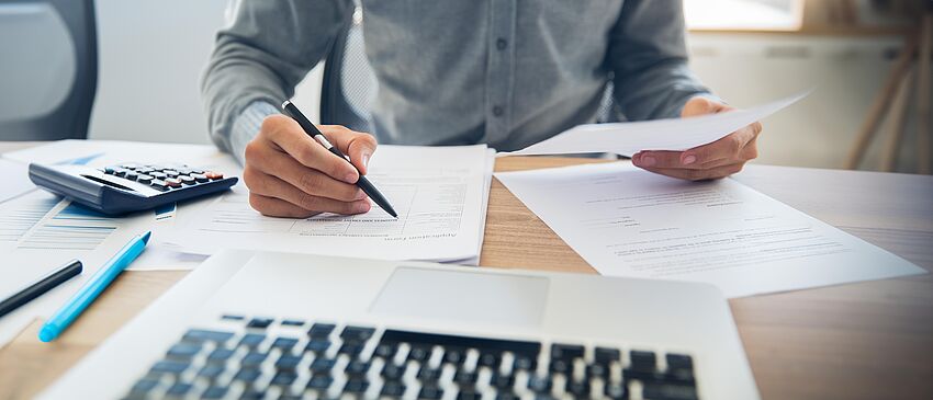 Eine Person sitz an einem Schreibtisch und prüft verschiedene Dokumente, welche er in der Hand hält bzw. auf dem Schreibtisch liegen. Vor ihm ein geöffneter Laptop.