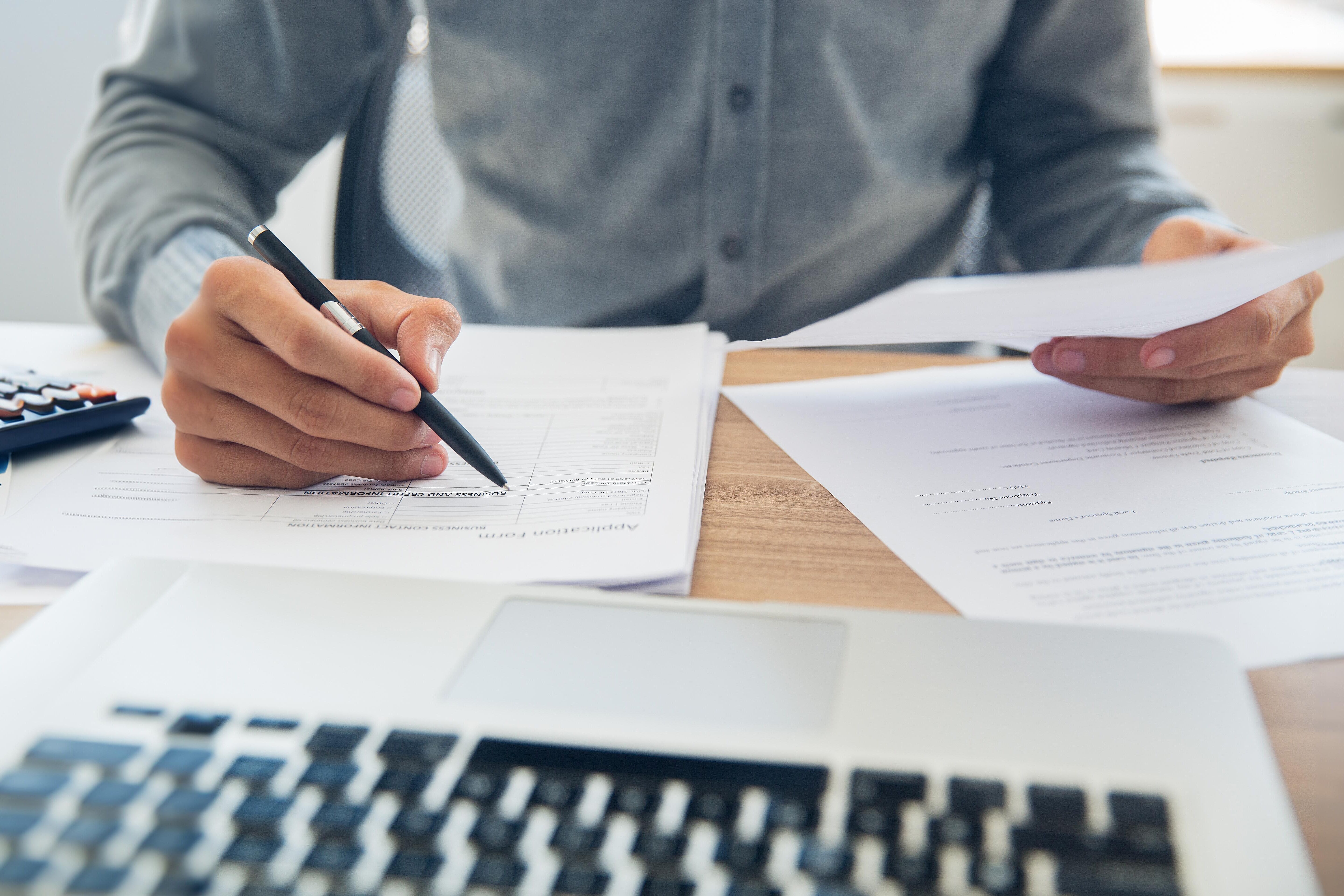 Eine Person sitz an einem Schreibtisch und prüft verschiedene Dokumente, welche er in der Hand hält bzw. auf dem Schreibtisch liegen. Vor ihm ein geöffneter Laptop.