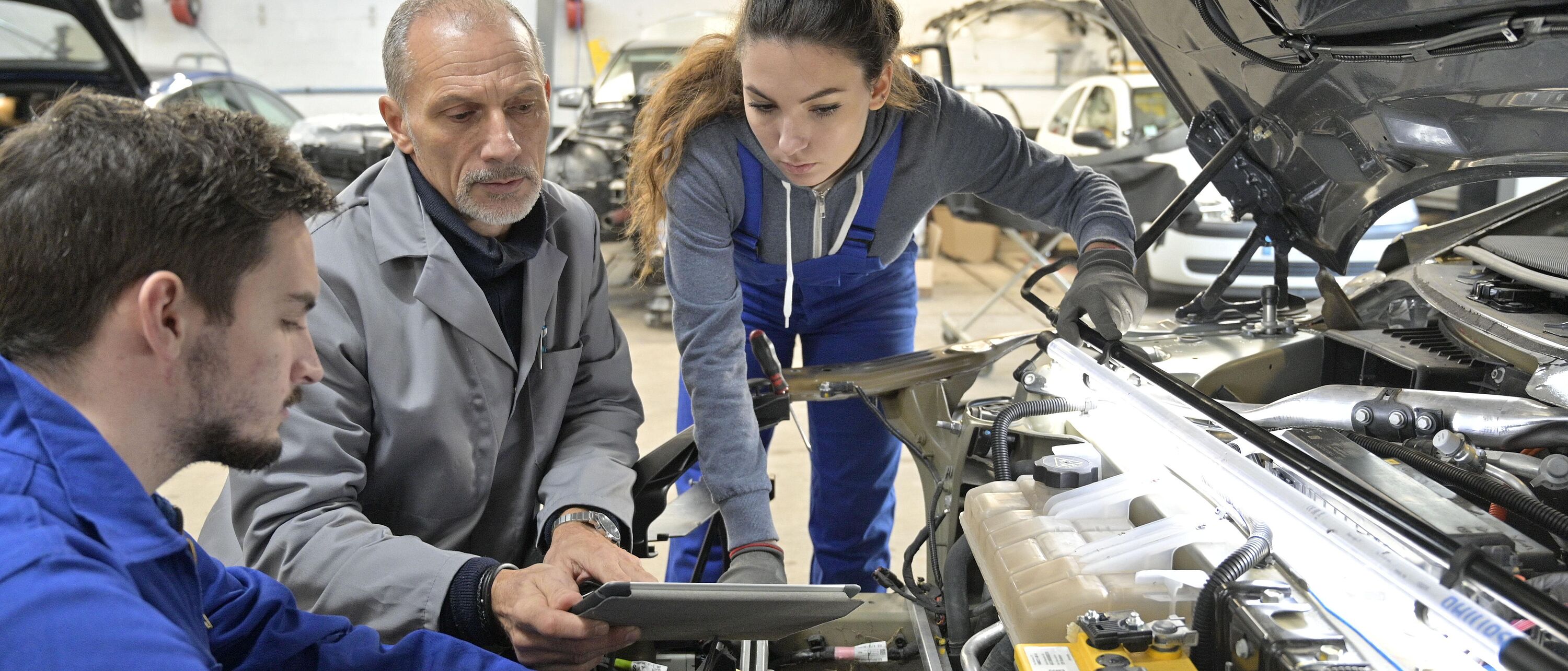 In einer Werkstatt sind drei Personen, ein Ausbilder mit zwei Auszubildenden vor einer offenen Motorhaube eines Fahrzeuges und führen Messungen mit Prüfgeräten aus.