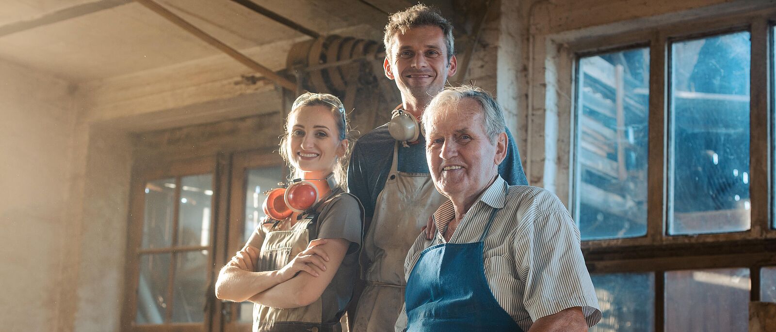3 Menschen, einer sitzend über 3 Generationen (älter, mittleren Altes und im Jugendalter) stehen in einer Werkstatt mit Fenster im Hintergrund.