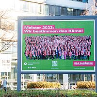 Eine große Plakatwand die vor dem Gebäude SLUB DrePunkt steht ist mit regionales Imagekampagnenmotiv beklebt. Darauf zu sehen sind allen Meisterabsolventen des Jahrganges 2023, darüber der Spruch \"Meister 2023: Wir bestimmen das Klima!\"