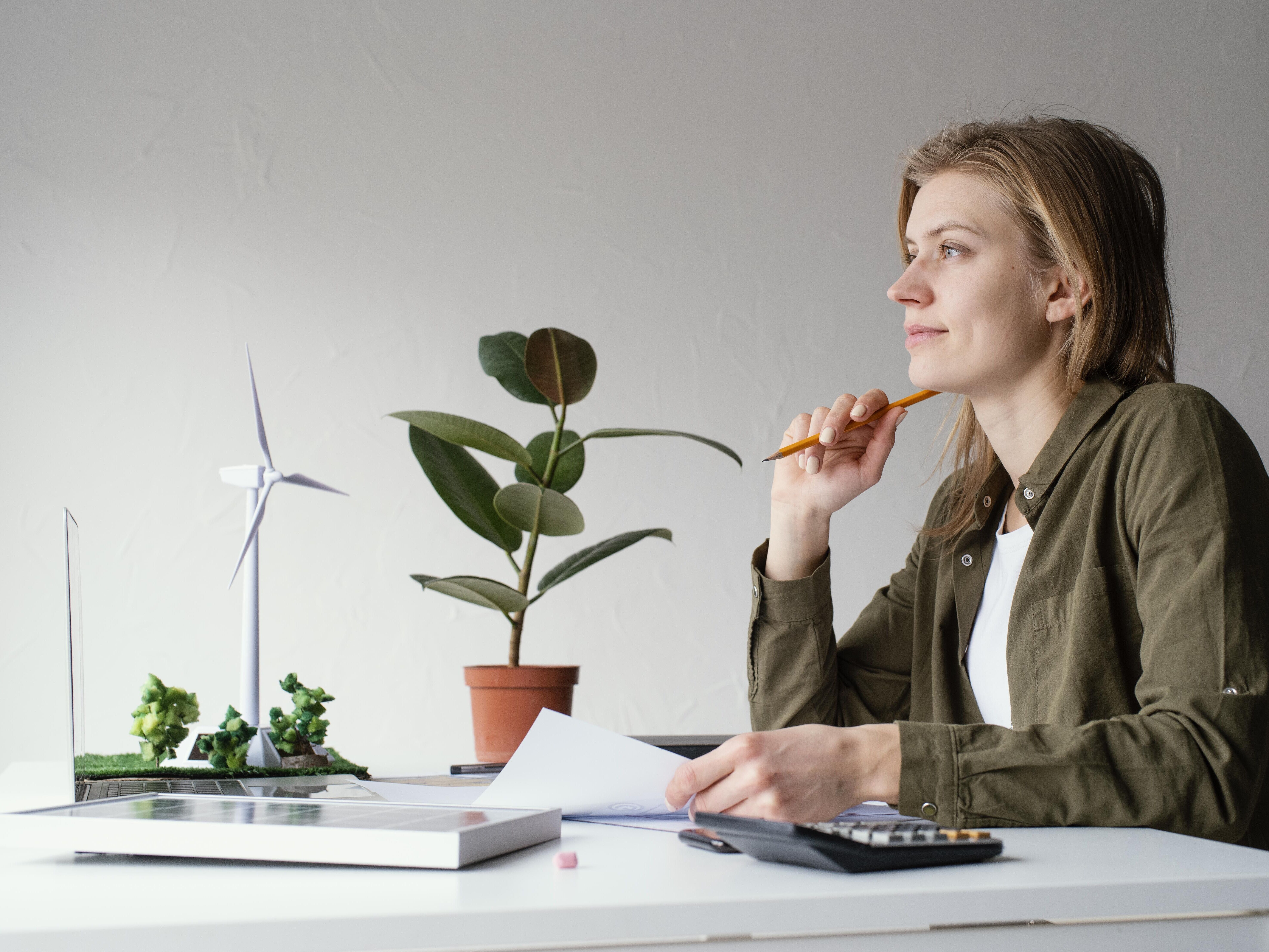 Eine Person sitz am Schreibtisch vor sich Unterlagen, in der rechten Hand ein Stift, den sie nachdenklich an das Gesicht hält. Auf dem Schreibtisch sind noch 3D Modelle von Windrädern, Photovoltaik und Solaranlagen sowie ein Laptop und eine Taschenrechner.