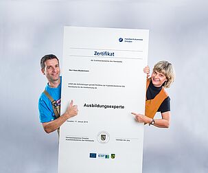 Zwei Ausbilder der Handwerkskammer Dresden stehen nebeneinander und halten mittig ein überdimensionales Zertifikat für Ausbilderexperten in den Händen.