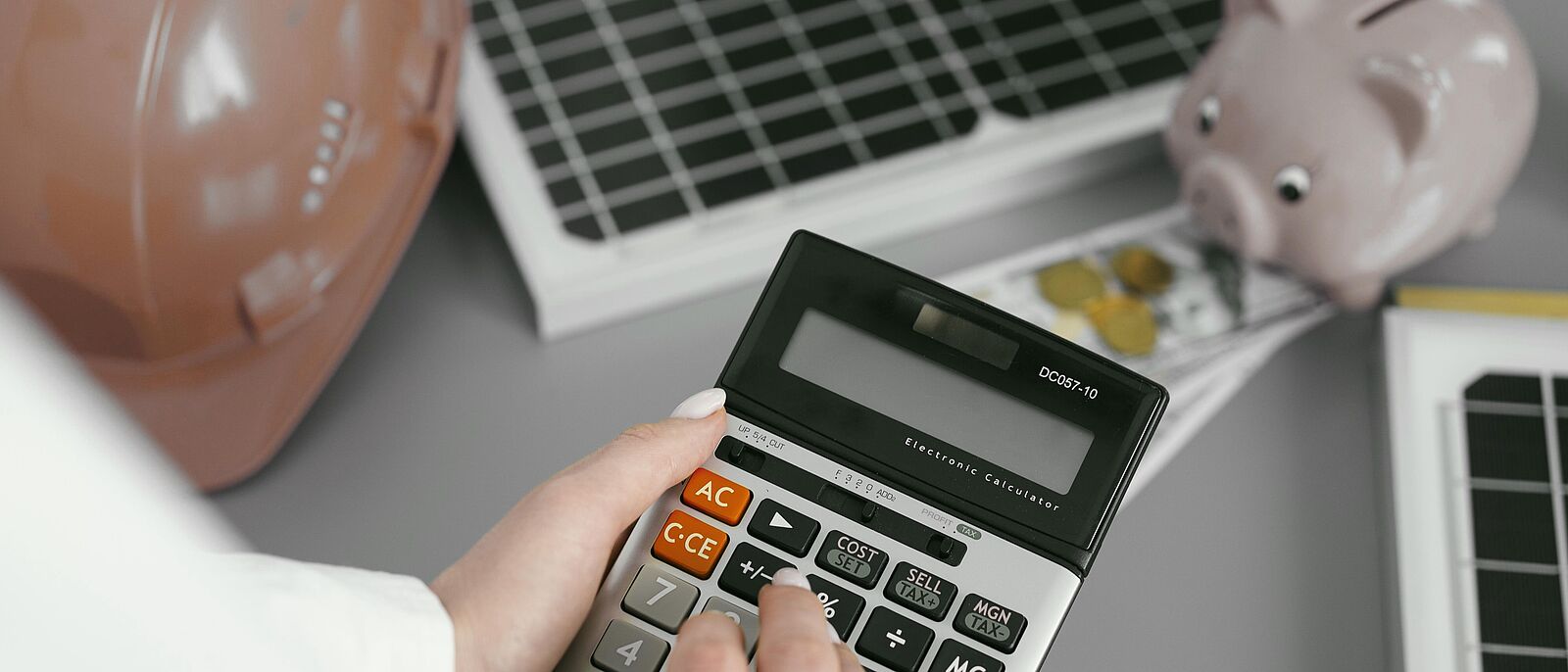 Makroaufnahme: Eine Person sitz an einem Büroplatz und führt Berechnungen an einem Taschenrechner aus. Im Hintergrund ein rosa Sparschwein, Münzen, Dokumente und die Tastatur  von einem PC.