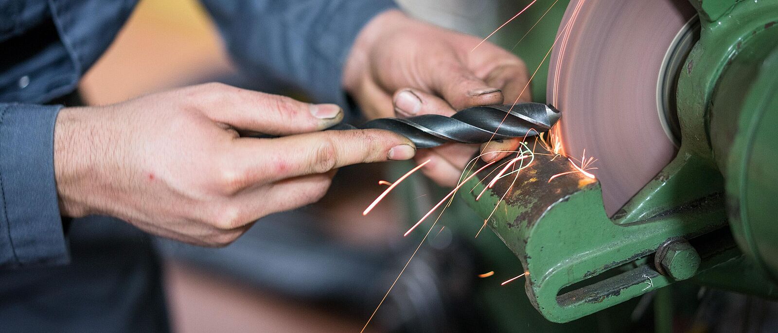 Eine Person in Arbeitskleidung eines Feinwerkmechanikers steht vor einer Werkbankmaschine und schleift ein Werkstück.