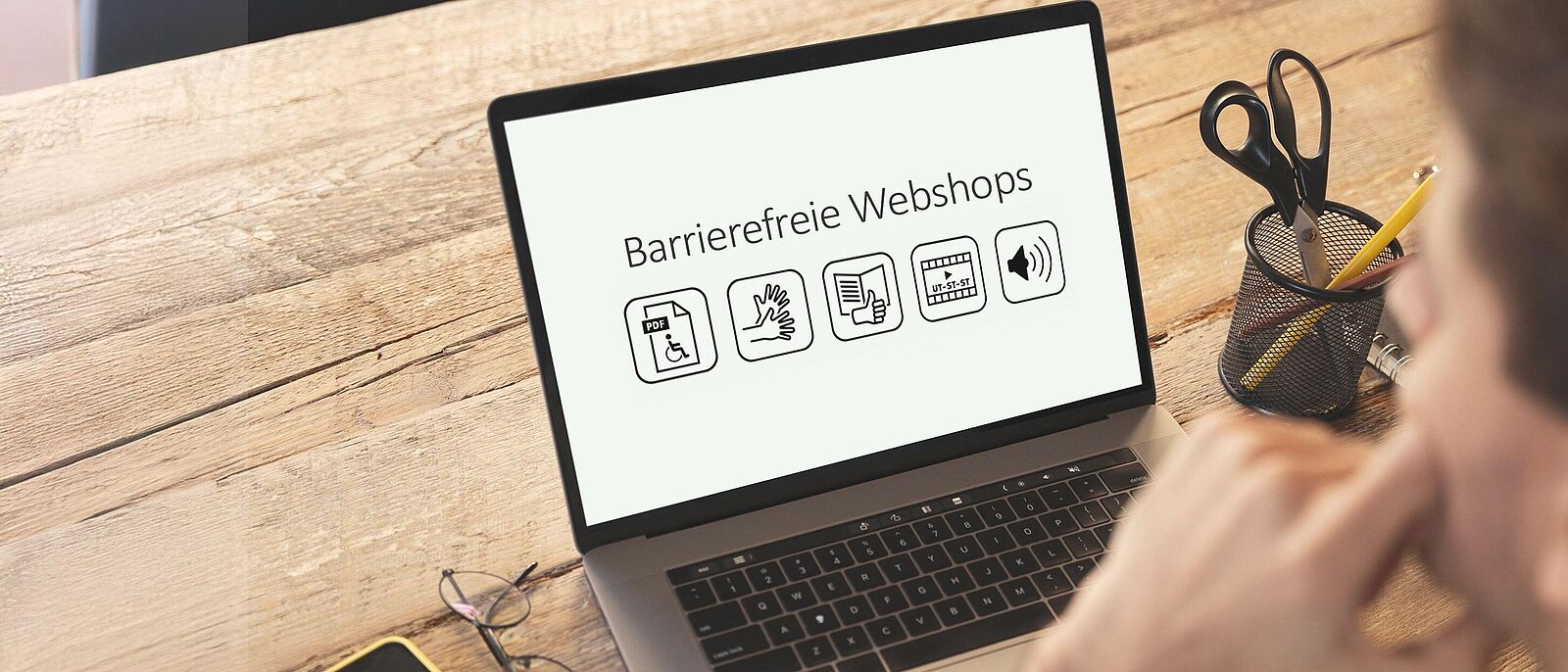 Eine Person sitzt vor einem Laptop und schreibt etwas darauf. Darüber grafische Elemente: ein Pfeil der von links kommend auf den Laptop zeigt. Auf dem Pfeil Icons zum Thema Barrierefreiheit.